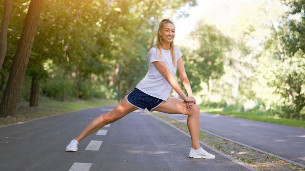 Woman runner stretching legs before exercising summer park morning Middle age athletic female warming up body before running Caucasian person warm up jogging Dressed white shirt shorts running track