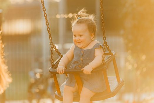 Toddler baby girl on a swing on the warm summer evening. Mother is swinging her young daughter on a sunny playground.
