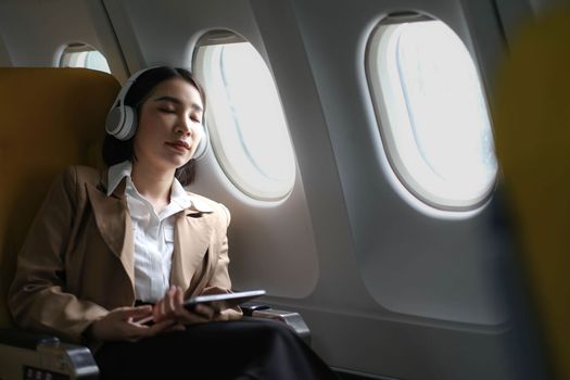 Young caucasian smiling female enjoying her comfortable flight while sitting in airplane cabin, listening to music in earphones and drinking water. Wifi internet access on board, passenger near window..