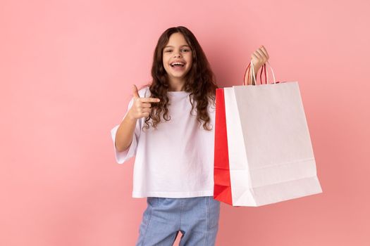 Portrait of excited little girl wearing white T-shirt pointing at shopping bags, looking at camera with pleased happy facial expression. Indoor studio shot isolated on pink background.