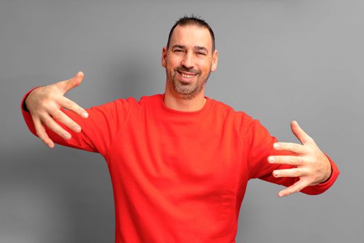 Handsome bearded man wearing casual red pullover looking at camera smiling with open arms for hugging. joyful expression embracing happiness. Isolated on gray background