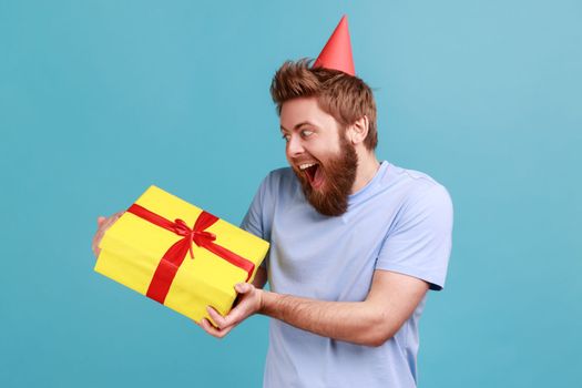 Portrait of extremely happy bearded man being very glad to get present from friend or girlfriend, holding gift box, expressing excitement. Indoor studio shot isolated on blue background.