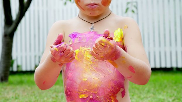 a little child, a four-year-old boy playing, painting with finger paints, decorating himself, in the garden, sitting on a blanket, on grass, lawn, in the summer. he's having fun. High quality photo