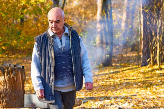 an outing or occasion that involves taking a packed meal to be eaten outdoors. Cheerful elderly man pensioner makes a fire for grilling at a picnic in autumn