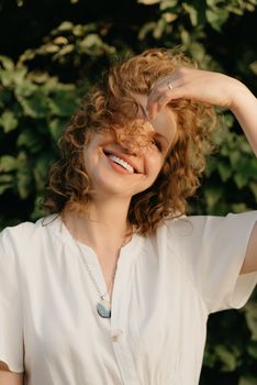 A smiling woman with curly hair is posing in the park in the evening. A close portrait of a lady in a white dress with green leaves in the background.