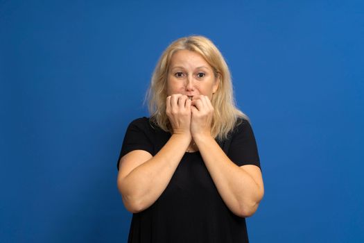 Caucasian blonde woman biting her nails out of fear, she is experiencing a dangerous and terrifying situation. Isolated on a blue studio background.