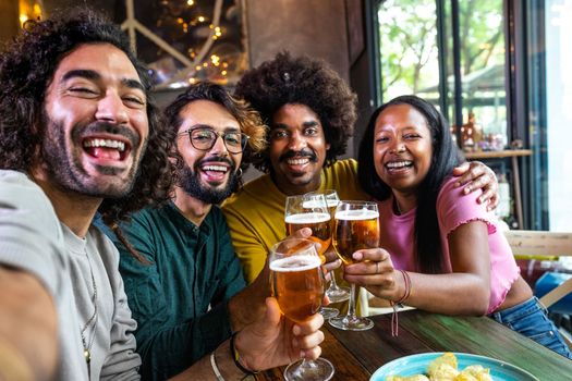 Happy multiracial friends in a bar toasting with beer taking selfie with phone looking at camera. Lifestyle concept.