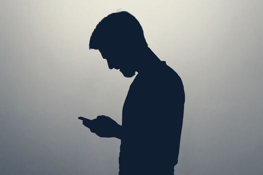 Male silhouette standing isolated on gray background looking at screen of smart phone, browsing web pages and chatting with friends.
