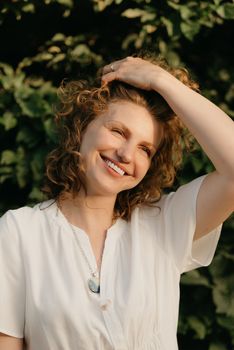 A smiling woman with curly hair is posing in the park in the evening. A lady in a white dress with her arm on her head with green leaves in the background.