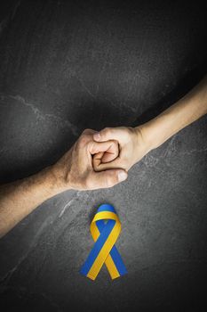 adult hand holding tight hand of child with yellow and blue ribbon on dark background. concept needs help and support, truth will win