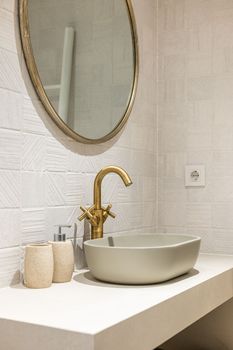 Oval stylish sink on white smooth marble countertop in modern bathroom. Copper-colored two-valve faucet blends harmoniously with round mirror hanging on wall and other objects in room