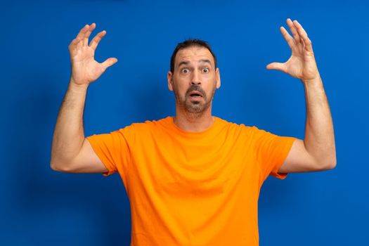 Hispanic man dreamy bearded melancholic surprised attractive man 40s in casual orange basic t-shirt spreading hands isolated on blue color background studio portrait. People lifestyle concept