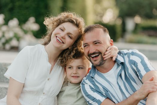 Father, mother, and son are sitting on the steps in the garden of an old European town. A close photo of a laughing family in the park in summer at sunset.