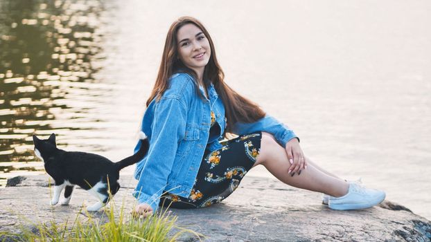 A girl sits on a rock with a cat against the background of a lake