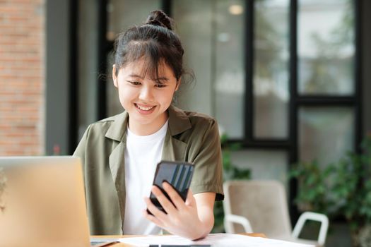 Young asian businesswoman uses a mobile phone to contact a customer, working hard at the office using laptop data graphs, planning for improvement, analyzing and strategizing for business growth. Business concept