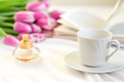 A still life with a white mug and a saucer, a golden bottle of perfume, an open book and a bouquet of beautiful pink tulips lies on a bed on a white fabric. close-up. copy space