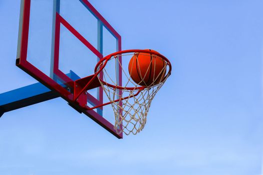 A basketball in a basket on an outdoor basketball court. A basketball is a goal in a basketball basket against the sky.
