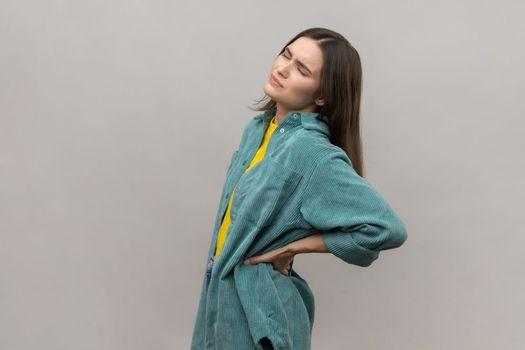 Portrait of frustrated anxious unhealthy woman holding hands on lower back, kidney injury, backache, wearing casual style jacket. Indoor studio shot isolated on gray background.