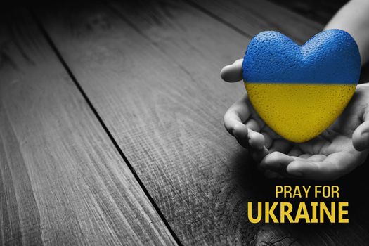 hand holds yellow and blue heart on dark wooden background with words pray for Ukraine. needs help and support, truth will win