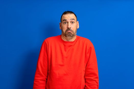 Frontal portrait of a bearded latino man in a red jumper doubting shrugging isolated on a blue studio background.