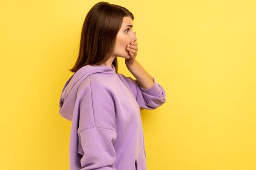 Side view portrait of shocked dark haired woman put hand on mouth and looking ahead, keeping terrible secret, wearing purple hoodie. Indoor studio shot isolated on yellow background.