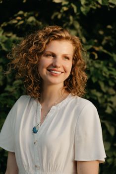 A smiling woman with curly hair is posing in the garden in the evening. A happy lady in a white dress with green leaves in the background.