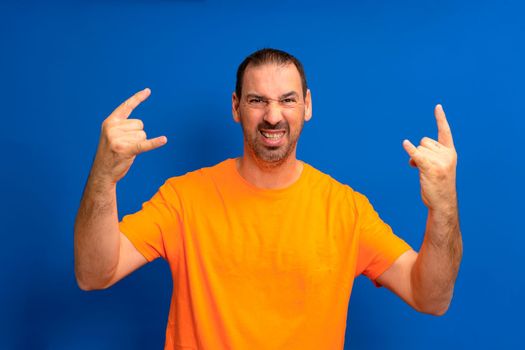 Bearded latino man wearing orange t-shirt with crazy expression doing rock symbol with hands up. Music star heavy concept