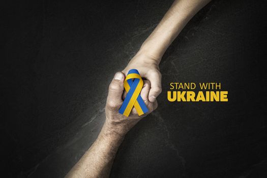 words stand with Ukraine and adult hand holding tight childs hand with yellow and blue ribbon. concept needs help and support