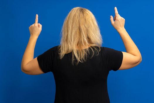 Caucasian blonde in a black T-shirt turned from behind and pointed with her index fingers, isolated against a blue background