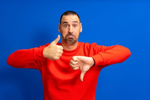 Photo of doubtful handsome bearded latino man wearing red jumper showing thumb up and down over isolated blue color background