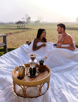 A couple of men and women at a cottage homestay in an outdoor bed in Northern Thailand Nan Province, couple waking up during sunrise in the rice fields in the morning drinking coffee outside