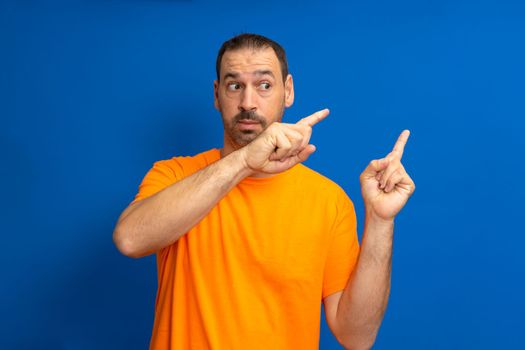 Bearded hispanic man wearing an orange t-shirt pointing to the side with index fingers isolated on a blue studio background
