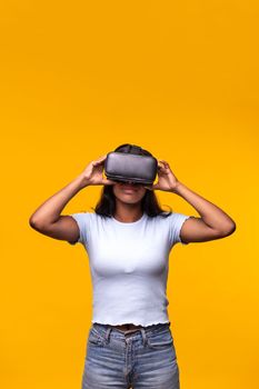 Young Indian woman wearing VR headset on yellow background. Vertical portrait of Asian female wearing virtual reality goggles. Technology.