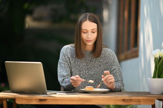 Millennial caucasian woman enjoy eating cake and using laptop at outdoor cafe.