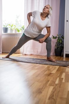 Domestic yoga practice. Positive mature lady standing in yoga pose, keeping balanced during covid lockdown at home. Happy senior lady in sportswear exercising