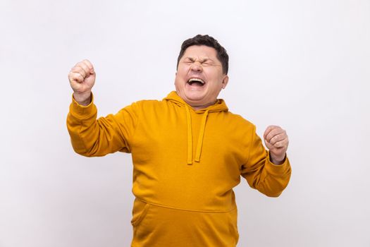Overjoyed handsome middle aged man expressing winning gesture with raised fists and screaming, celebrating victory, wearing urban style hoodie. Indoor studio shot isolated on white background.