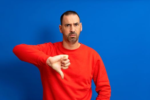 Bearded hispanic man wearing red sweater sulking shows thumb down with negative attitude, isolated over blue background