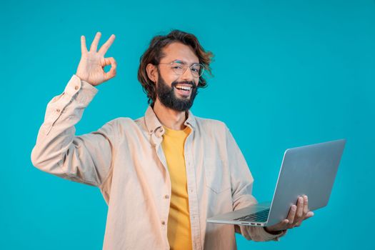 Portrait of a man holding laptop computer and celebrating success isolated over blue background. High quality photo