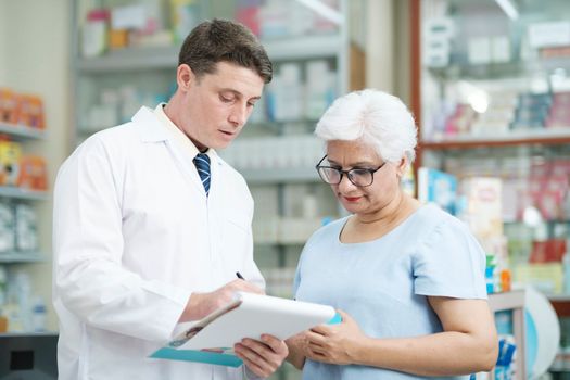 Smart male pharmacist at the drugstore wearing white gown talking, giving advice, explaining, suggesting, and recommending to client or patient about the prescription and medications. Medicine and healthcare concept.