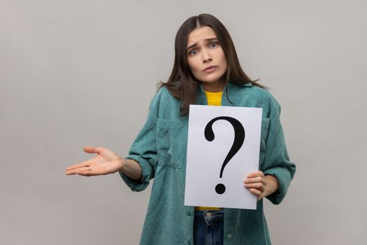 Portrait of confused puzzled woman looking at camera, holding paper with question mark, thinks about tasks, wearing casual style jacket. Indoor studio shot isolated on gray background.