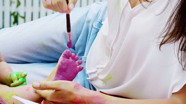 a young beautiful mother and her one-year-old daughter playing with finger paints in the garden, sitting on a plaid, a blanket, a coverlet, on the grass, lawn, in the summer. High quality photo