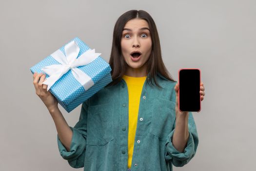 Astonished woman holding gift box and cell phone with empty display for online shopping advertising, expressing shock, wearing casual style jacket. Indoor studio shot isolated on gray background.