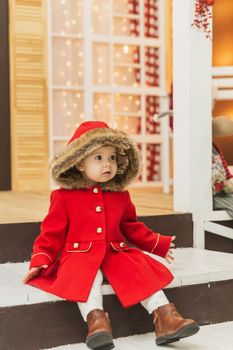 charming baby in a red coat with a hood sits on the porch.