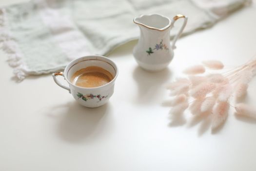 White cup with coffee and milk jug on a white wooden background, closeup. Energy breakfast, morning routine concept. coffee cup and milk pitcher on white table background