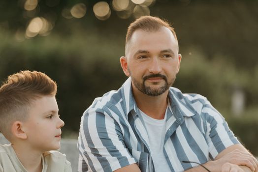 Father and son are sitting on the steps in the garden of an old European town. A close photo of a little boy and his dad in the park in summer at sunset.