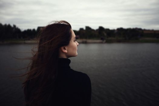 cute teenager girl wind inflates luggage enjoys fresh air by the water in the evening outdoors dressed in black gloomy clothes looks into the distance. High quality photo