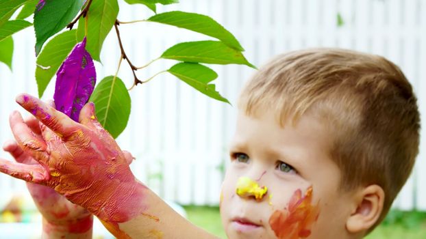 a little child, a four-year-old boy playing, painting with finger paints, decorating leaves on the trees in the garden, in the summer. High quality photo