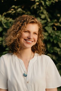 A smiling woman with curly hair is posing in the park at the sunset. A close portrait of a lady in a white dress with green leaves in the background.