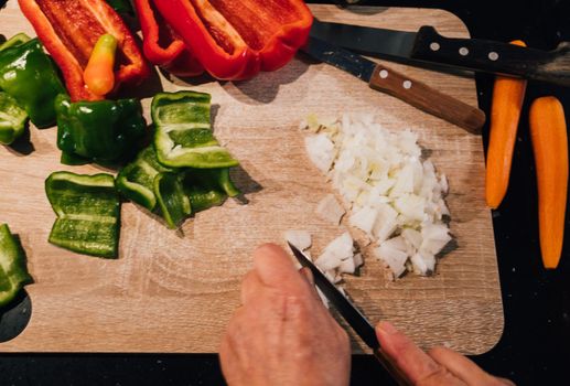 top view of woman hands cutting onion on wooden board with colourful red , green , orange and white vegetables around.