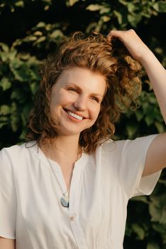 A smiling woman with curly hair is posing in the park in the evening. A happy lady in a white dress with her arm on her head with green leaves in the background.